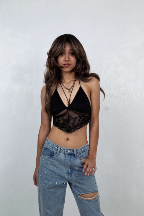 Portrait of Dallas M. smiling while wearing a black lace halter top and rhinestone-studded jeans against a white studio wall.