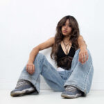 Portrait of Dallas M. seated on the floor wearing a black lace halter top, oversized jeans, and sneakers in a minimalist studio setting.