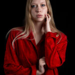 Close-up portrait of TamLynn Link wearing a red blouse while gently touching her face against a black background.