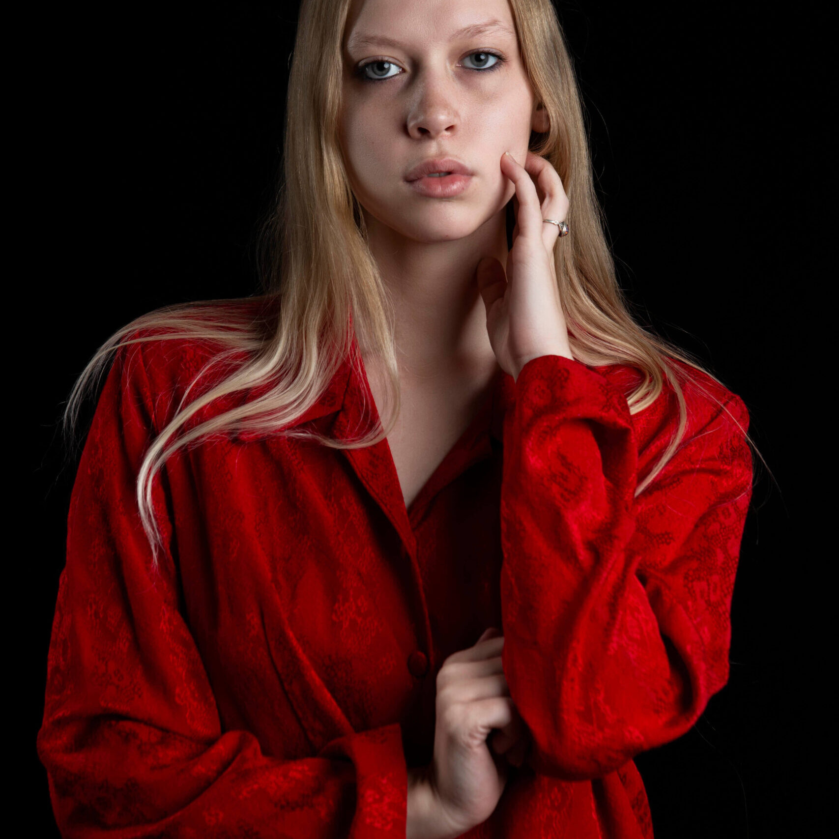 Close-up portrait of TamLynn Link wearing a red blouse while gently touching her face against a black background.