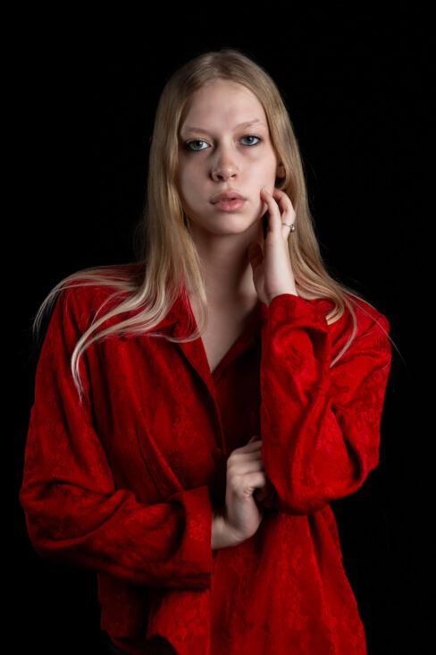 Close-up portrait of TamLynn Link wearing a red blouse while gently touching her face against a black background.