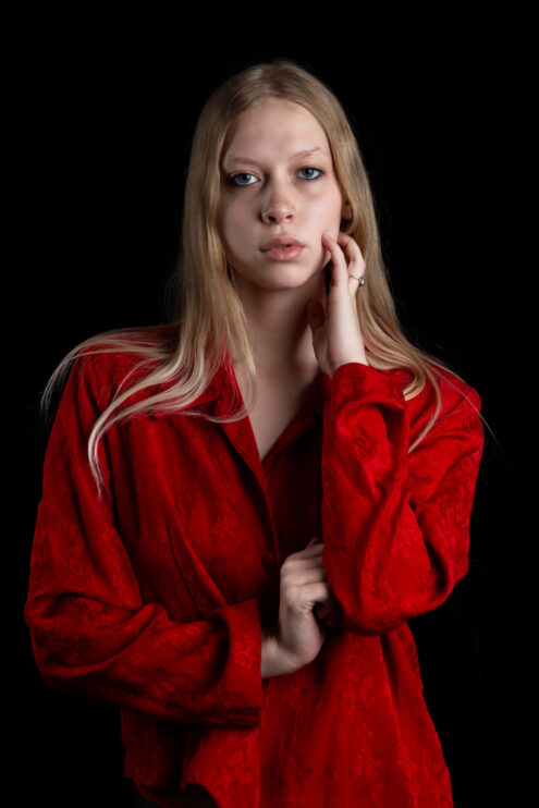 Close-up portrait of TamLynn Link wearing a red blouse while gently touching her face against a black background.