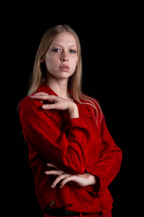 Portrait of TamLynn Link wearing a red blouse with a graceful hand pose against a black studio background.