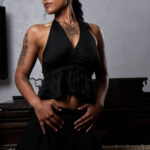 Portrait of Vinny Young standing in front of a grand piano wearing a black halter top and skirt in a studio setting.