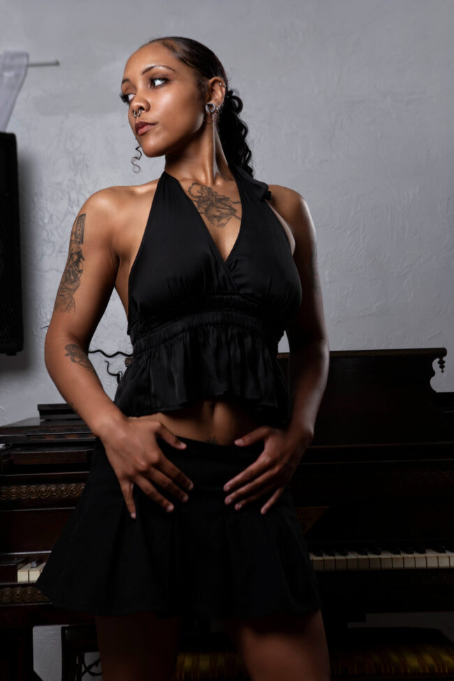Portrait of Vinny Young standing in front of a grand piano wearing a black halter top and skirt in a studio setting.