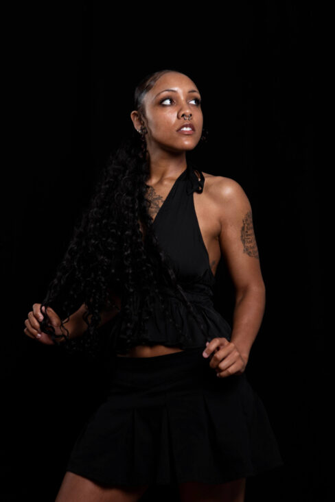 Studio portrait of Vinny Young with long curly hair wearing a black outfit against a dark background.