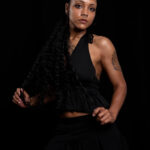 Portrait of Vinny Young wearing a black outfit and long curly hair against a dark studio background.