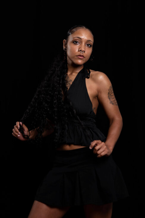 Portrait of Vinny Young wearing a black outfit and long curly hair against a dark studio background.