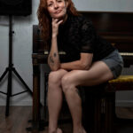 Thegoddessek seated on a piano bench, resting her chin on her hand while posing in a black sheer top and gray shorts in a softly lit studio.