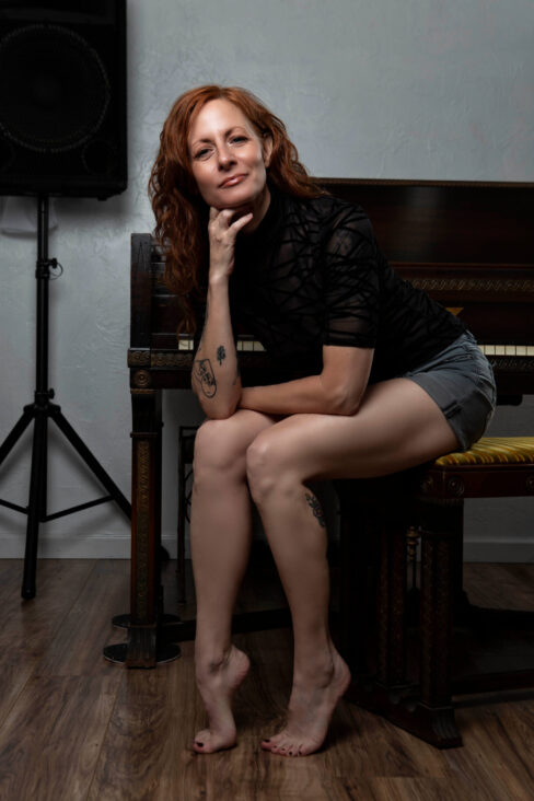Thegoddessek seated on a piano bench, resting her chin on her hand while posing in a black sheer top and gray shorts in a softly lit studio.