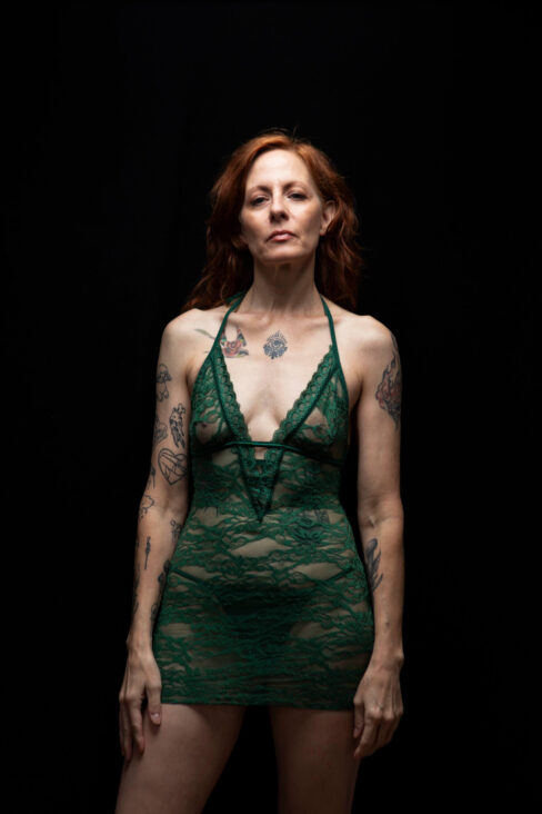 Thegoddessek standing against a black backdrop wearing a green lace dress, looking directly at the camera in a dramatic studio portrait.