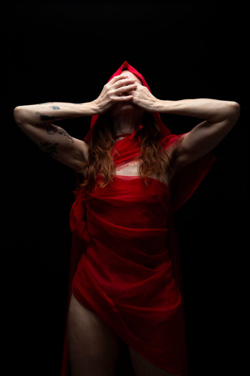 Thegoddessek draped in flowing red fabric with a hood covering part of her face, posing dramatically against a black background.