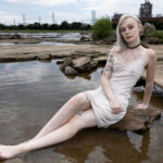 Portrait of a model seated on riverside rocks beside a calm pool of water, wearing a light dress in a rugged natural landscape.