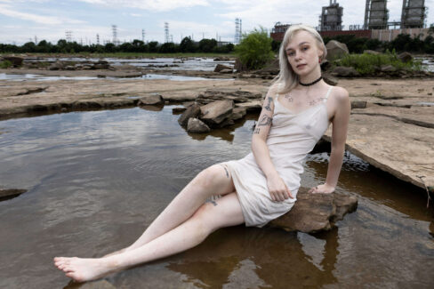 Portrait of a model seated on riverside rocks beside a calm pool of water, wearing a light dress in a rugged natural landscape.