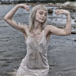 Model kneeling in shallow water with eyes closed and arms raised gracefully, wearing a wet dress stained with mud.