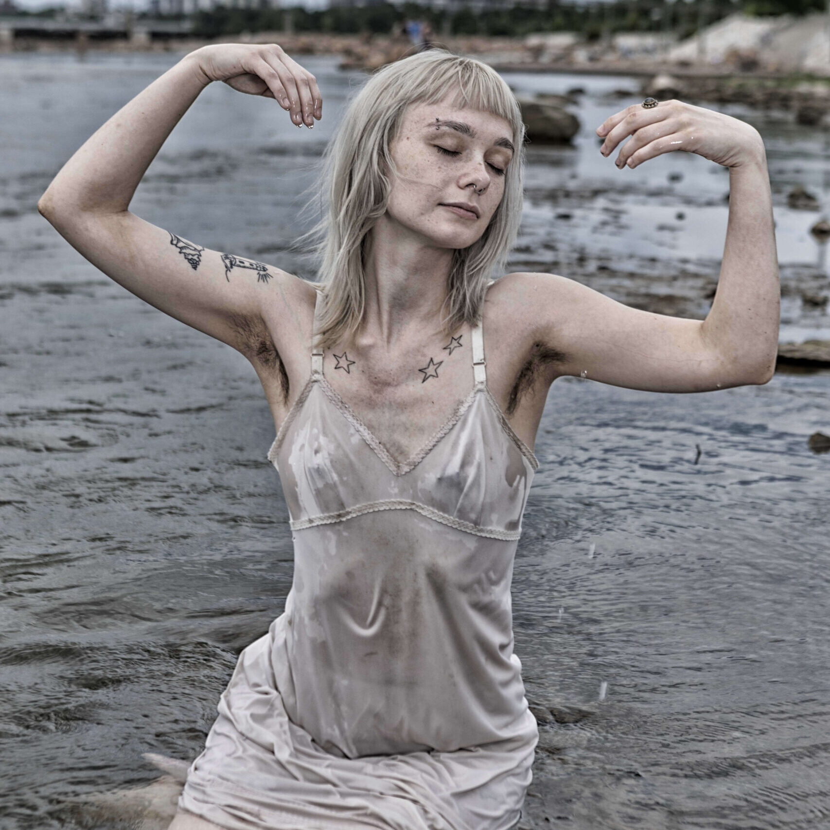Model kneeling in shallow water with eyes closed and arms raised gracefully, wearing a wet dress stained with mud.