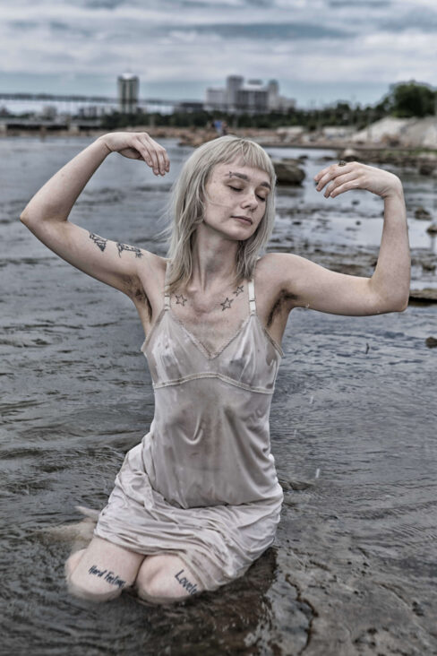 Model kneeling in shallow water with eyes closed and arms raised gracefully, wearing a wet dress stained with mud.