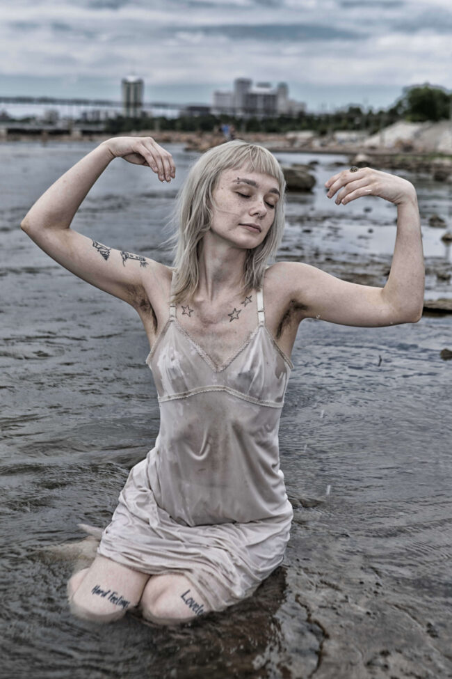 Model kneeling in shallow water with eyes closed and arms raised gracefully, wearing a wet dress stained with mud.