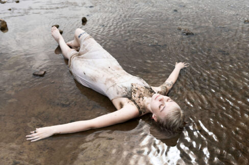 Model lying in a shallow river wearing a light-colored dress covered in mud, arms extended as water flows around her.