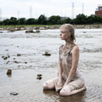 Model kneeling in shallow water with a mud-covered dress, looking toward the horizon with an industrial riverside landscape in the background.