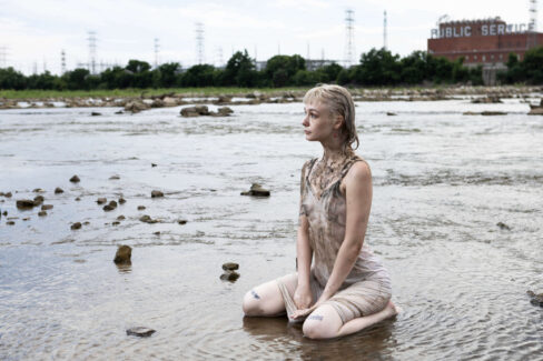 Model kneeling in shallow water with a mud-covered dress, looking toward the horizon with an industrial riverside landscape in the background.