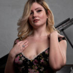 Close-up portrait of Molly Must Die wearing a black floral dress while posing beside a grand piano in a studio setting.