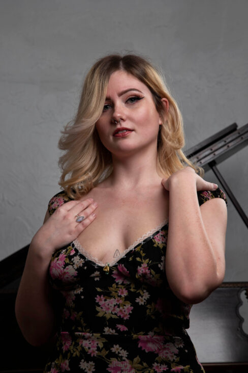 Close-up portrait of Molly Must Die wearing a black floral dress while posing beside a grand piano in a studio setting.