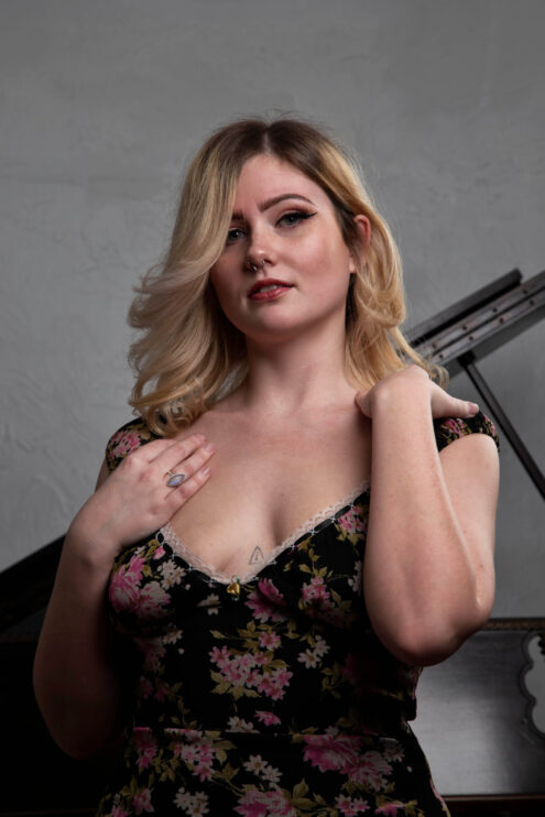 Close-up portrait of Molly Must Die wearing a black floral dress while posing beside a grand piano in a studio setting.