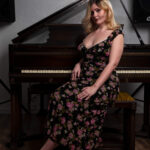 Full-length portrait of Molly Must Die seated on a piano bench wearing a floral dress in front of a grand piano.