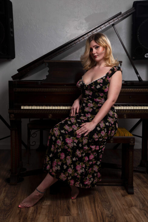 Full-length portrait of Molly Must Die seated on a piano bench wearing a floral dress in front of a grand piano.