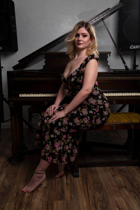 Portrait of Molly Must Die seated sideways on a piano bench wearing a floral dress in a studio.