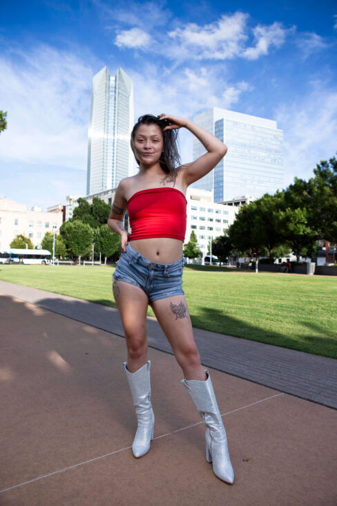 Augustine Amasaman posing in front of the Oklahoma City skyline wearing a red tube top, denim shorts, and white boots.