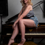Lifestyle portrait of Molly Must Die sitting on a piano bench wearing a gingham top and denim shorts in a studio setting.