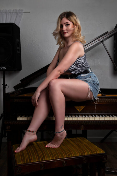 Lifestyle portrait of Molly Must Die sitting on a piano bench wearing a gingham top and denim shorts in a studio setting.