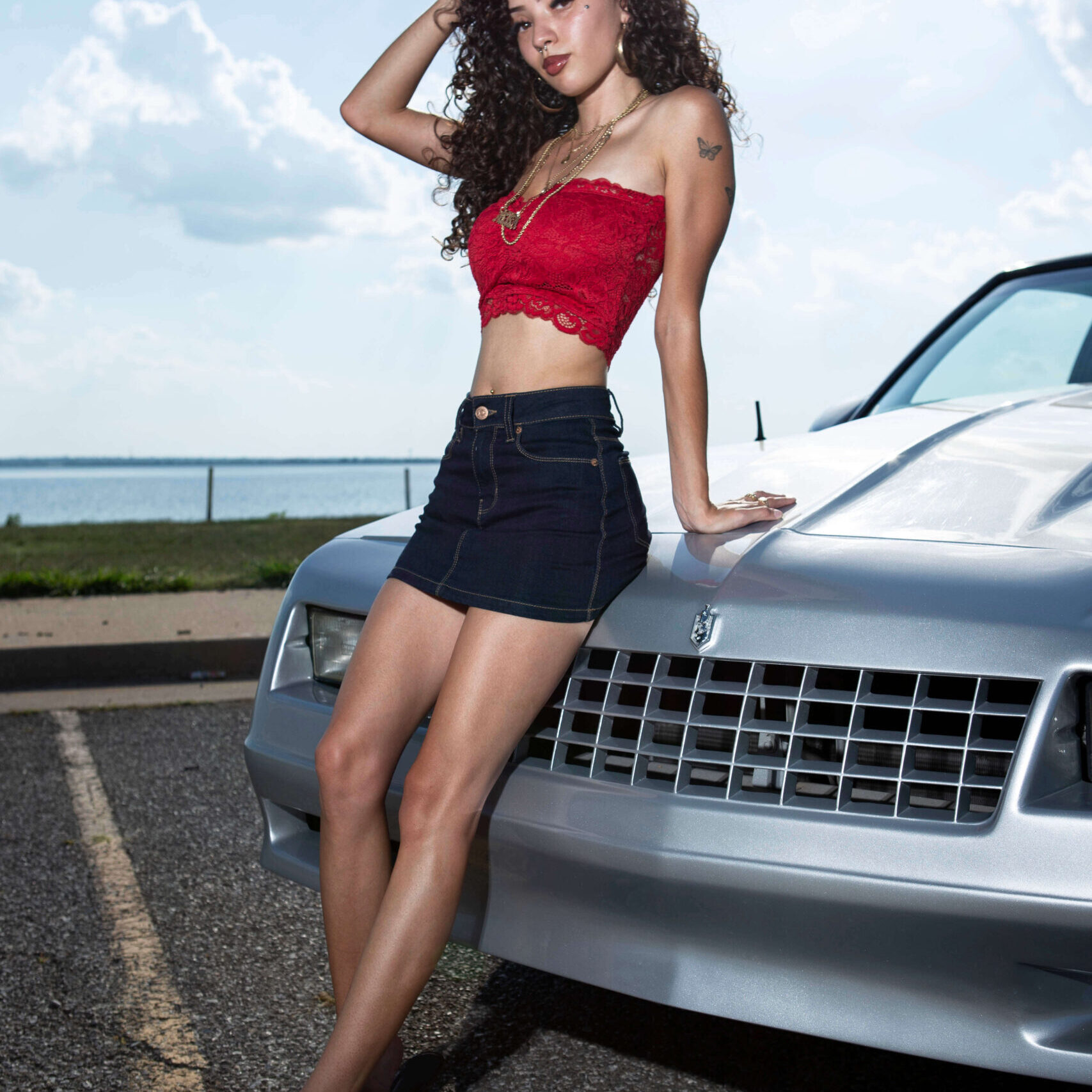 Jas posing beside a silver Chevrolet Monte Carlo near a lakeside roadway while wearing a red lace top and denim skirt.