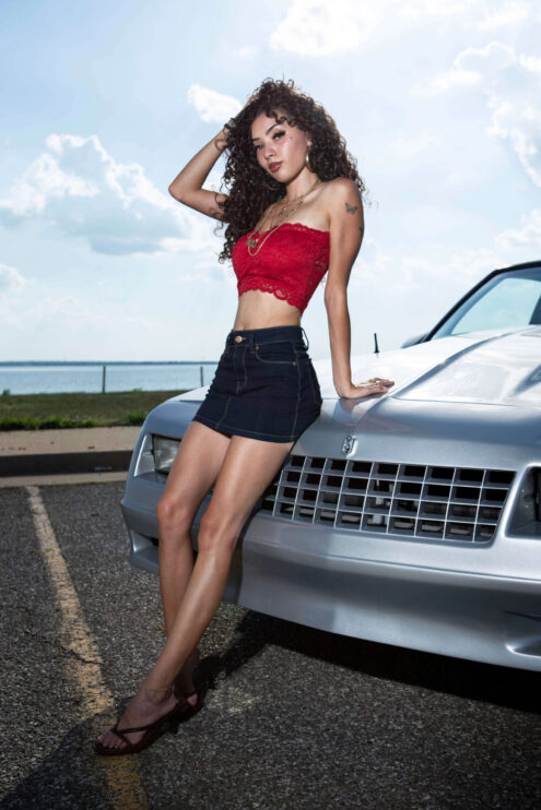 Jas posing beside a silver Chevrolet Monte Carlo near a lakeside roadway while wearing a red lace top and denim skirt.