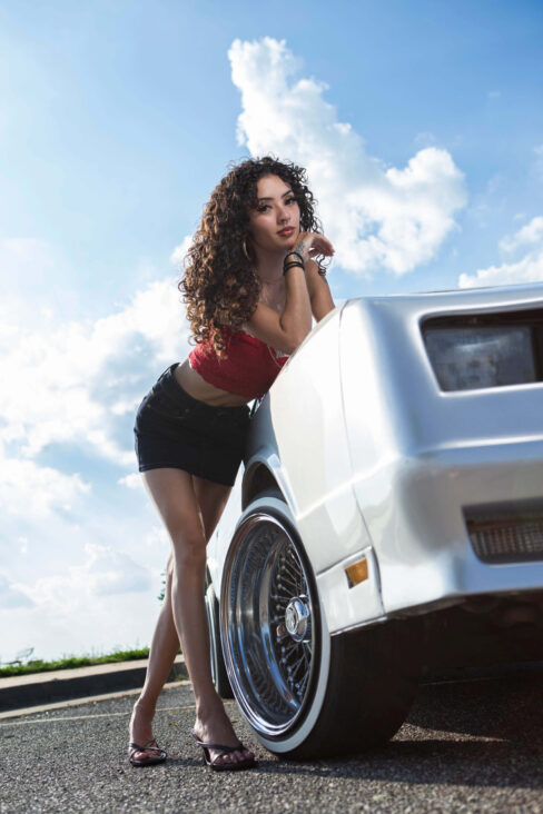 Jas leaning against the front fender of a silver Monte Carlo beneath a bright summer sky.