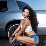 Jas crouching beside a silver Monte Carlo while wearing a white crop top, denim shorts, and white heels.