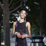 Bratzdoll smiling beneath the trees near Devon Tower in downtown Oklahoma City wearing a black and purple fitted dress during an outdoor portrait session.