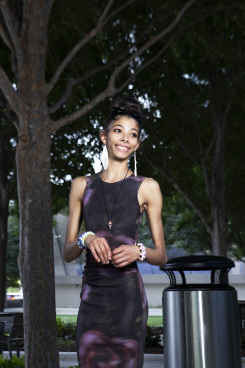 Bratzdoll smiling beneath the trees near Devon Tower in downtown Oklahoma City wearing a black and purple fitted dress during an outdoor portrait session.