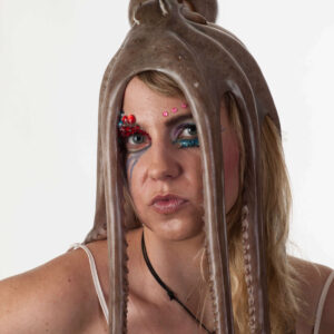 Anne Alexandra poses against a white studio background with an octopus draped over her head, its long tentacles framing her face. Colorful theatrical eye makeup, decorative gemstones and delicate white straps complete her unusual bridal-inspired appearance.