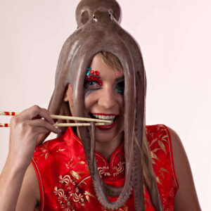 Anne Alexandra smiles while wearing an octopus on her head and holding chopsticks near her mouth. She wears a vibrant red Chinese-inspired dress with gold embroidery and colorful theatrical makeup against a pale studio background.