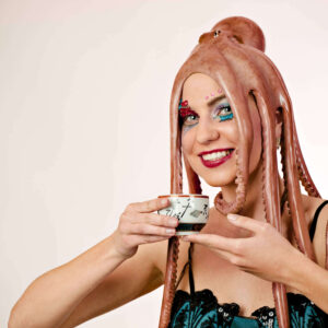 Anne Alexandra wearing an octopus as a headdress, colorful theatrical makeup and a black and turquoise outfit while holding a decorative teacup against a white studio background.