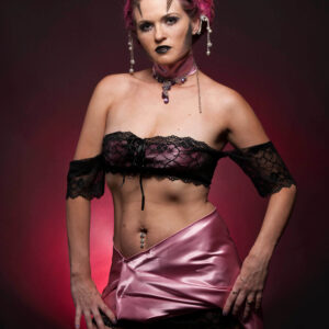 Traci Lacek poses in an elaborate alternative fashion look featuring magenta braided hair, geometric face makeup, black lace, a pink satin skirt, and jeweled accessories.