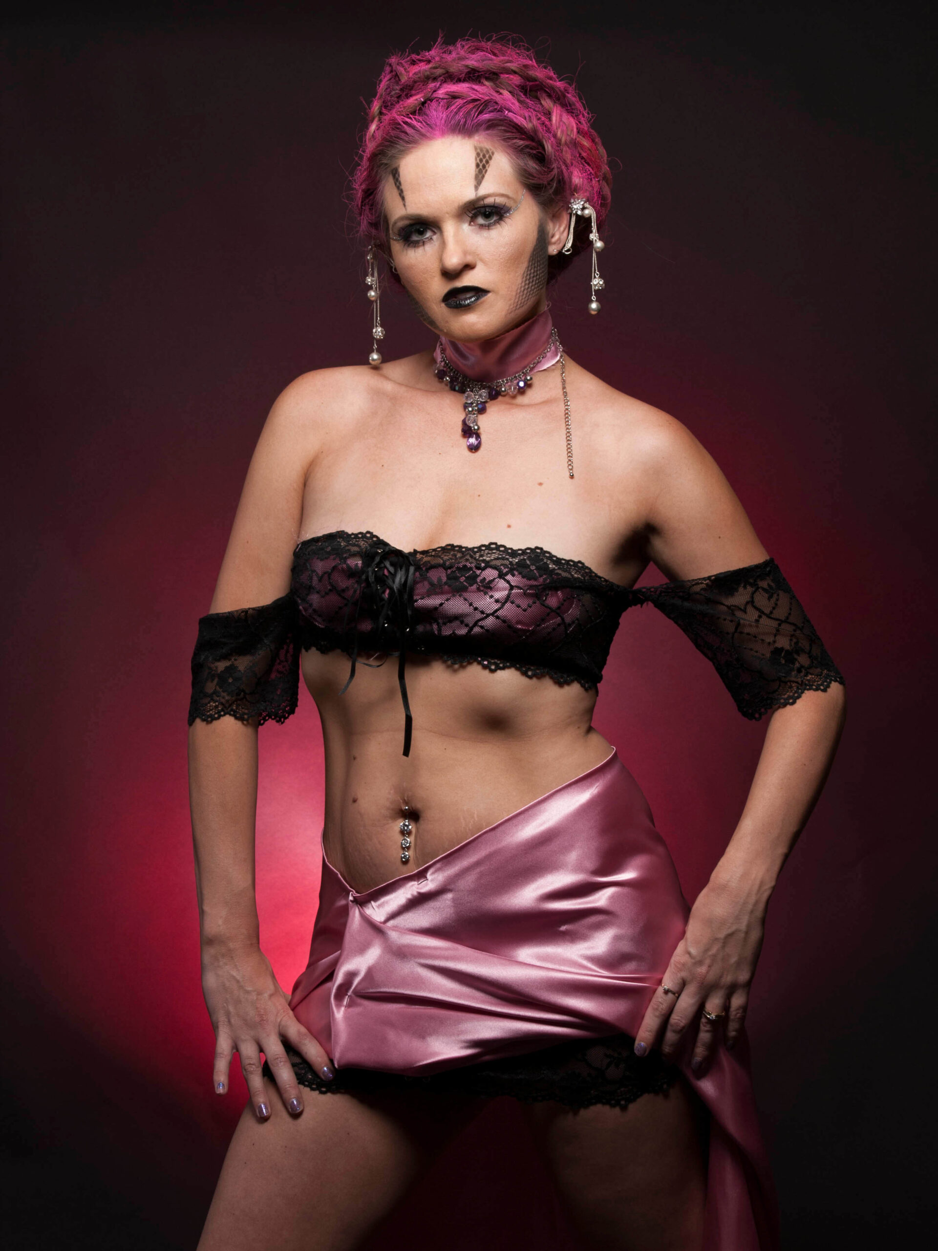 Traci Lacek poses in an elaborate alternative fashion look featuring magenta braided hair, geometric face makeup, black lace, a pink satin skirt, and jeweled accessories.