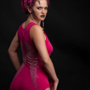 Traci Lacek poses against a black studio background wearing a vivid pink fitted outfit with rhinestone detailing, elaborate magenta braided hair, and geometric fantasy makeup.
