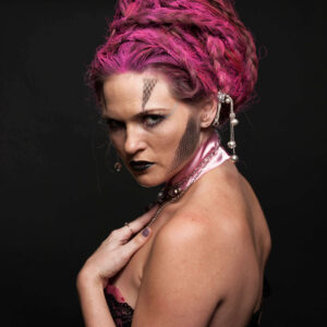 Traci Lacek poses in profile against a black background with an elaborate magenta braided updo, geometric face makeup, black lipstick, jeweled hair accessories, and black lace styling.