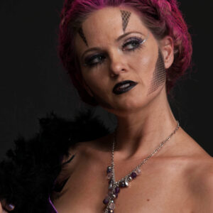 Close-up portrait of Traci Lacek with magenta-styled hair, dramatic geometric face makeup, dark lipstick, purple lingerie, and a jeweled necklace against a dark background.