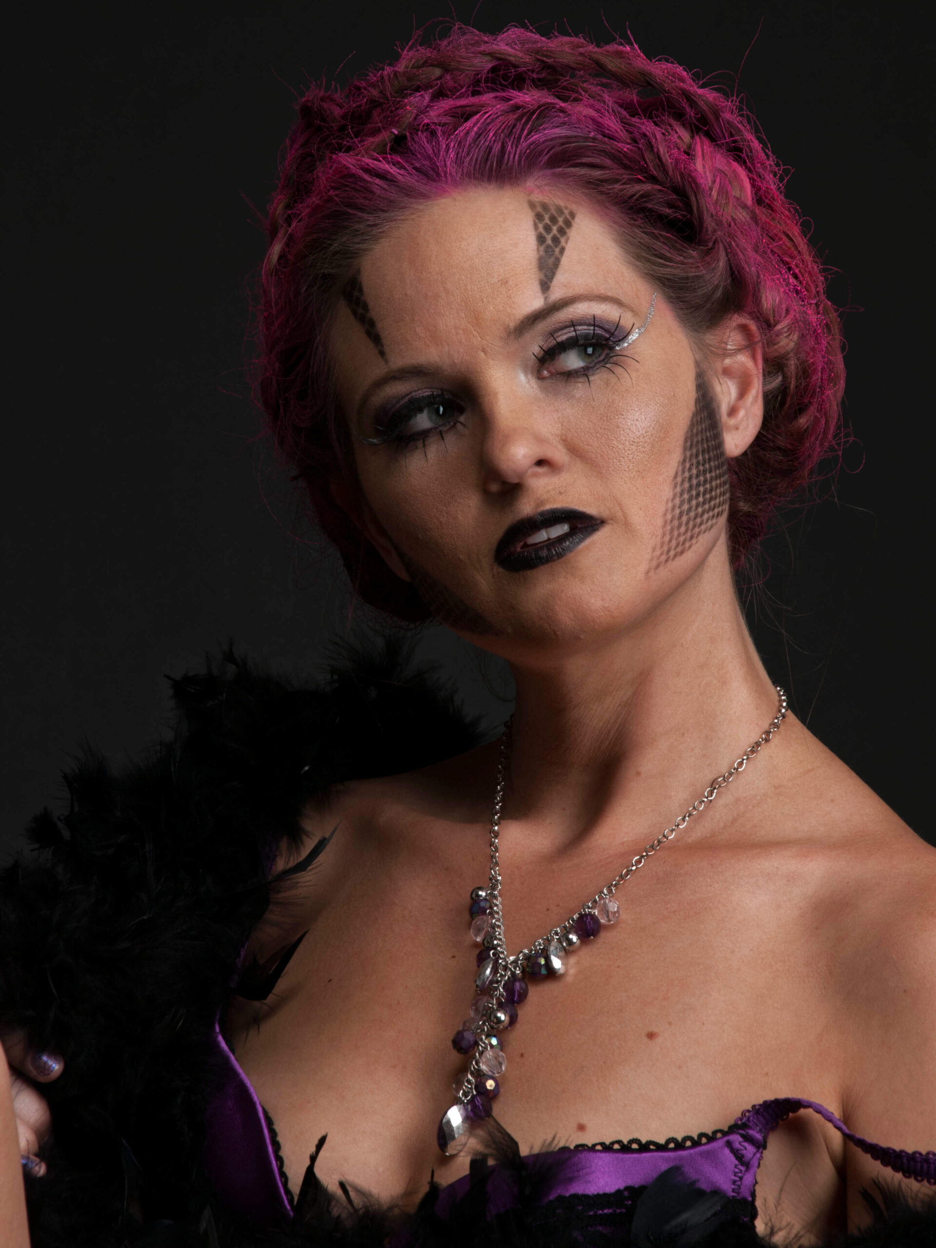 Close-up portrait of Traci Lacek with magenta-styled hair, dramatic geometric face makeup, dark lipstick, purple lingerie, and a jeweled necklace against a dark background.