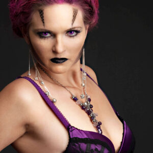 Close-up portrait of Traci Lacek with elaborate magenta braided hair, geometric face makeup, vivid purple eyes, black lipstick, and purple-and-black lingerie.